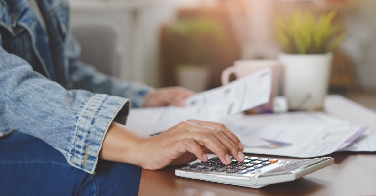 A woman in a jean jacket reaching out to a calculator on a desk while holding a document in her other hand.